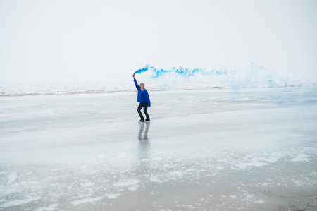 A caucasian woman is skating on a frozen lake holding colored blue smoke in her handsの写真素材