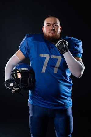 An Asian man with a red beard in a blue american football uniform holds a helmet on a black backgroundの写真素材