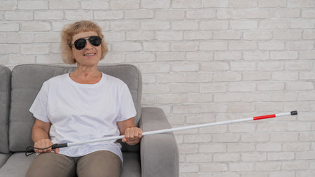 An elderly blind woman wearing sunglasses and with a tactile cane sits on the sofaの写真素材