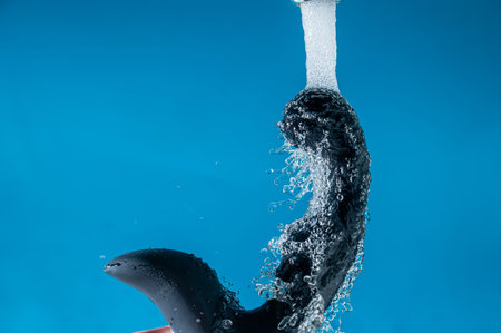 A woman washes a black prostate stimulator under running water on a blue background. Sex toy hygiene conceptの写真素材
