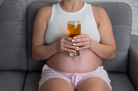 Close-up of the belly of a pregnant woman holding a glass of beer while sitting on the sofaの写真素材