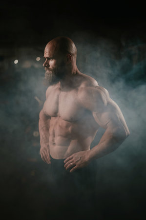 A muscular bald man poses shirtless in the dark amid smoke. A bodybuilder shows off his form in the gymの写真素材