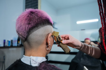 The hairdresser shaves the temple of a female client. Rear view of a woman with short pink hair in a...