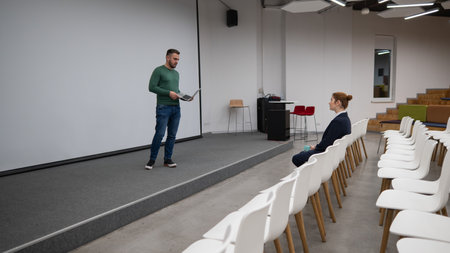 A red-haired Caucasian businesswoman sits in the front row of an empty conference room. Bearded man...