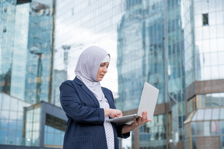 Thoughtful business woman in hijab and suit is holding a laptop outdoorsの写真素材