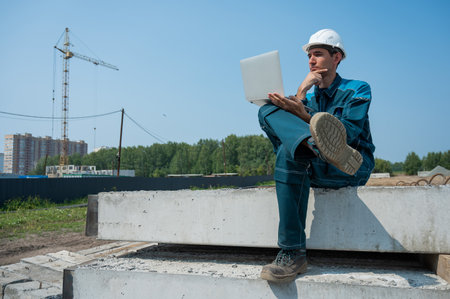 Caucasian male builder in hardhat sits on floor slabs and uses laptop at construction site