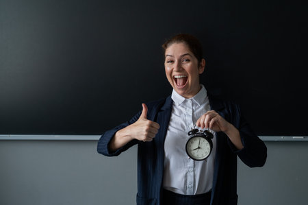 Happy female teacher in pantsuit holding an alarm clock standing at the school board and showing thumbs upの写真素材
