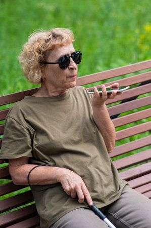 An elderly blind woman sits on a bench in the park and talks on a smartphone. Vertical photoの写真素材