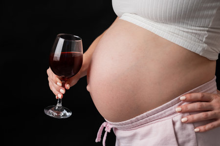 Close-up of the belly of a pregnant woman holding a glass of red wine while sitting on the sofaの写真素材