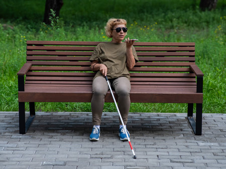 An elderly blind woman sits on a bench in the park and talks on a smartphone