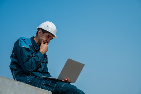 Caucasian male builder in hardhat sits on floor slabs and uses laptop at construction siteの写真素材