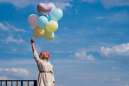 A woman with colored hair with an armful of balloons against a blue skyの写真素材