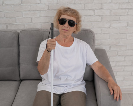 An elderly blind woman wearing sunglasses and with a tactile cane sits on the sofaの写真素材