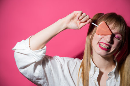 Portrait of a young woman with braces and bright makeup eating a lollipop on a pink backgroundの写真素材
