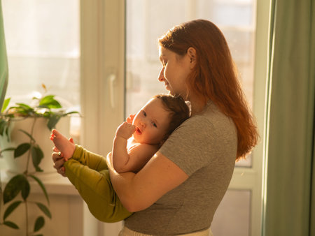 Caucasian woman tenderly holds her newborn son while standing near the windowの写真素材