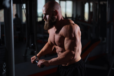 A bearded, barechested man in shorts is working out at the gym, using a machine to strengthen his...