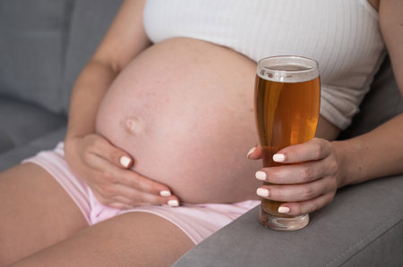 Close-up of the belly of a pregnant woman holding a glass of beer while sitting on the sofaの写真素材
