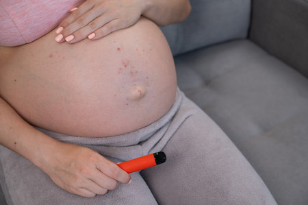 A pregnant woman smokes a vape while sitting on the couch. Close-up of the belly with allergiesの写真素材