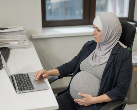 Pregnant Caucasian woman in hijab working at a computer in the officeの写真素材