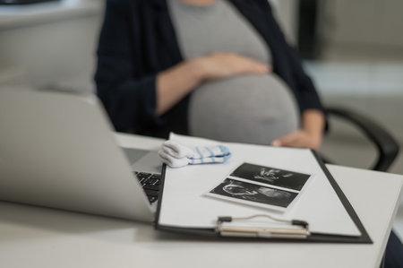 A pregnant woman works on a laptop in the office and looks at a photo from an ultrasound scan of the fetus. Belly close-upの写真素材