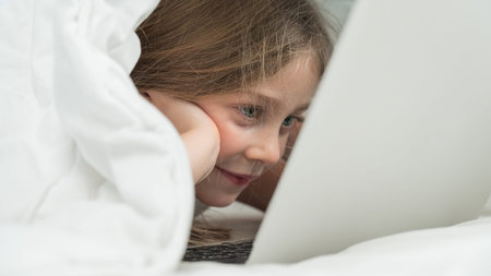 Portrait of a cute little girl using a laptop while lying under a blanketの写真素材