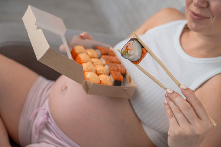 A pregnant woman sits on the sofa and eats rolls from a box. Food deliveryの写真素材