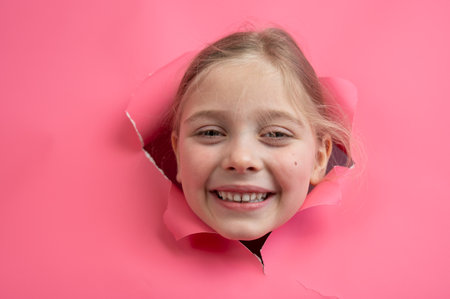 Cute Caucasian girl peeks out of a hole in a paper pink backgroundの写真素材