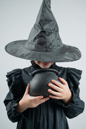 Portrait of a little Caucasian girl in a witch costume holding a cauldron on a white background. Vertical photoの写真素材