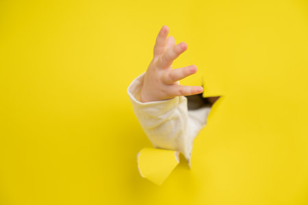 Babys hand sticks out through a hole into a yellow cardboard backgroundの写真素材