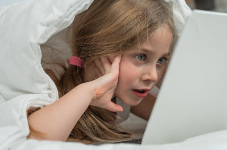 Portrait of a cute little girl using a laptop while lying under a blanketの写真素材
