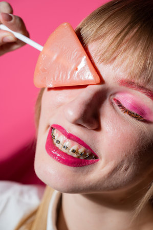 Portrait of a young woman with braces and bright makeup eating a lollipop on a pink background. Vertical photoの写真素材