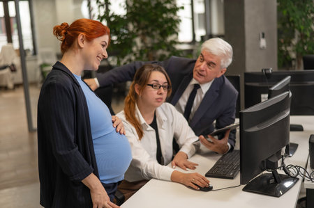 An elderly man, a Caucasian woman and a pregnant woman are discussing work issues at the computerの写真素材