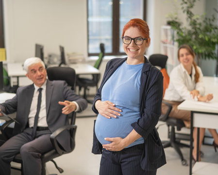 A happy pregnant woman stands in the middle of the office next to a Caucasian woman and an elderly man working at computersの写真素材