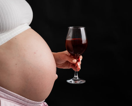 Close-up of the belly of a pregnant woman holding a glass of red wine while sitting on the sofa. Copy spaceの写真素材