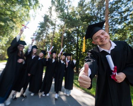 Happy young caucasian male graduate showing thumbs up. A group of graduate students outdoorsの写真素材