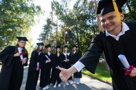 Happy young caucasian male graduate showing thumbs up. A group of graduate students outdoorsの写真素材