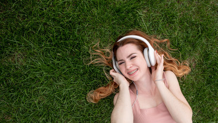 Top view of a young red-haired woman lying on the grass and listening to music on headphonesの写真素材