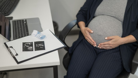 A pregnant woman works on a laptop in the office and looks at a photo from an ultrasound scan of the fetus. Belly close-upの写真素材