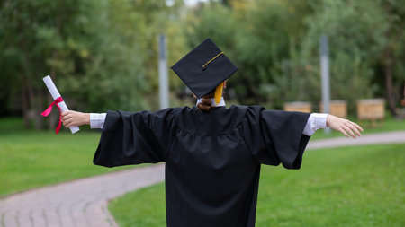Back view of happy caucasian woman in graduate gown holding diploma outdoorsの写真素材