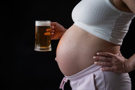 Close-up of the belly of a pregnant woman holding a glass of beer on a black background. Skin rashの写真素材