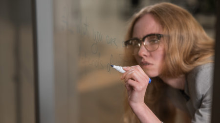 Caucasian woman with glasses writes text in English on a glass wall