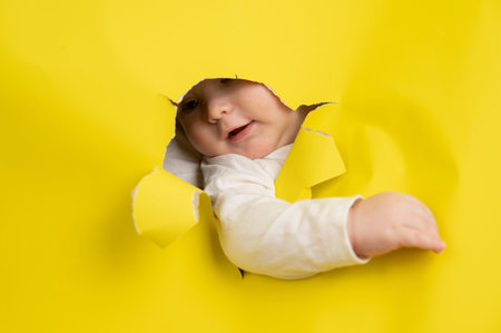 Cute Caucasian baby sticking out of a hole in a paper yellow backgroundの写真素材