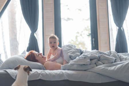 Red-haired Caucasian woman lies in bed with her baby son in a country house with panoramic windowsの写真素材