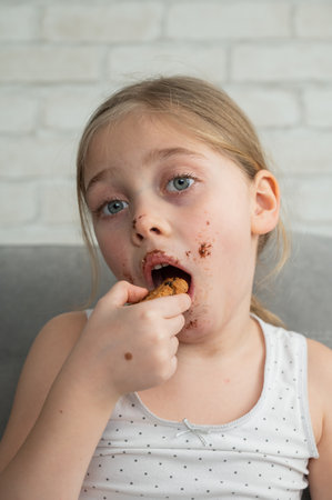 A cute little girl covered in chocolate eats cookies while sitting on the sofa. Vertical photoの写真素材