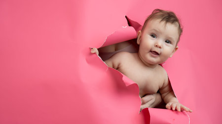 Cute Caucasian newborn baby boy peeks out of a hole in a paper pink background
