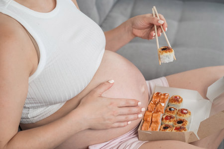 A pregnant woman sits on the sofa and eats rolls from a box. Food delivery. Close up of bellyの写真素材