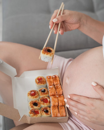 A pregnant woman sits on the sofa and eats rolls from a box. Food delivery. Close up of bellyの写真素材