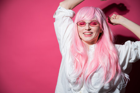 Close-up portrait of a young woman with braces in a pink wig and sunglasses on a pink background. Copy spaceの写真素材