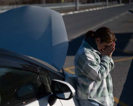 Sad Caucasian woman calling on phone while standing near car with open hoodの写真素材