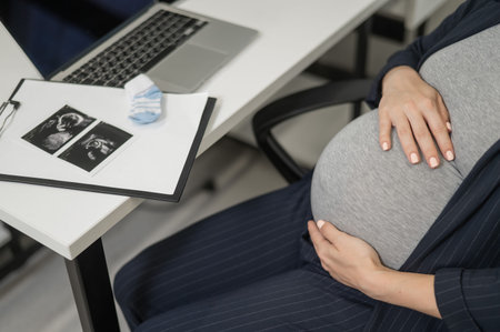 A pregnant woman works on a laptop in the office and looks at a photo from an ultrasound scan of the...